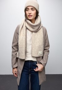 Cappello e sciarpa in maglia beige con una texture morbida, avvolti attorno a un maglione chiaro e a un cappotto grigio. Jeans in denim scuro completano l'outfit.