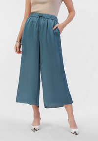 Teal wide-leg trousers with an elastic waistband, drawstring, side pockets, and smooth texture; worn with white pointed-toe heels.