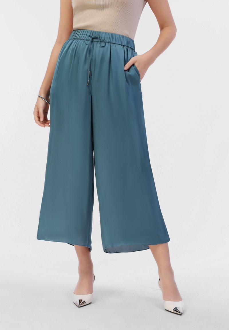 Teal wide-leg trousers with an elastic waistband, drawstring, side pockets, and smooth texture; worn with white pointed-toe heels.