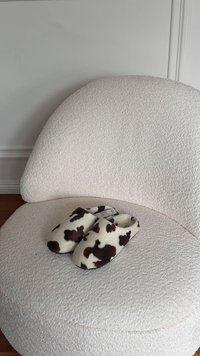 Pantofole con stampa a motivo mucca, con una texture morbida e soffice, caratterizzate da una base bianca e macchie marrone scuro, posate su una sedia chiara con superficie texturizzata.