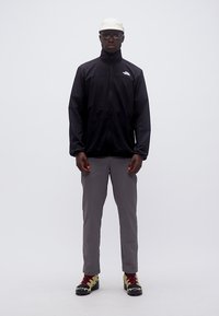 Άνδρας που φοράει μαύρο μπουφάν North Face, γκρι παντελόνι, μπεζ καπέλο, σκούρα γυαλιά ηλίου και παπούτσια πεζοπορίας, στέκεται μπροστά σε απλό γκρι φόντο.