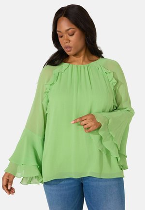 Grüne Chiffon-Bluse mit Rüschendetails an den Ärmeln und am Ausschnitt sowie einem lockeren, fließenden Schnitt. Kombiniert mit blauen Jeans.