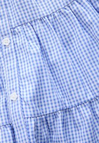 Mėlynos ir baltos spalvų languota medžiaga su tekstūruotu paviršiumi turi sagas ir surinktas detales, sukuriant sluoksniuotą išvaizdą.