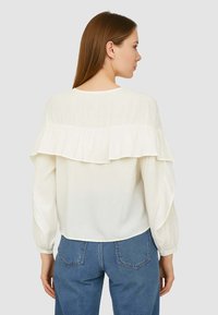 Blusa bianca con maniche lunghe a sbuffo, caratterizzata da un ruffles a strati sul retro. Tessuto morbido e leggero, abbinato a jeans blu.