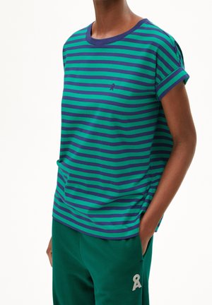 Person iført en grøn og mørkeblå stribet kortærmet skjorte og grønne bukser med et hvidt nøglehul-formet logo på låret.