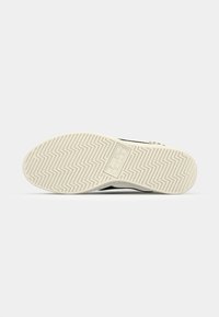 Sneaker sole with a textured, zigzag pattern in cream color, featuring a logo embossed in the center. Rubber material offering grip.