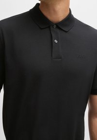 Svart polo skjorta tillverkad av texturerat tyg, med en plaggknäppning med två knappar, en klassisk krage och en diskret logotyp på vänster bröst.