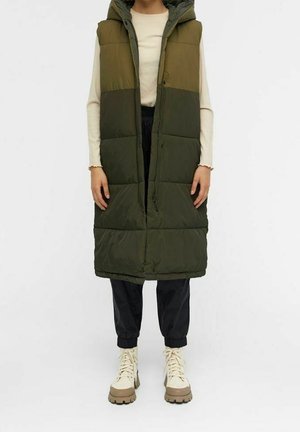 Personne portant un long gilet matelassé sans manches vert olive sur un haut crème, un pantalon noir et des bottines beiges à lacets, debout.