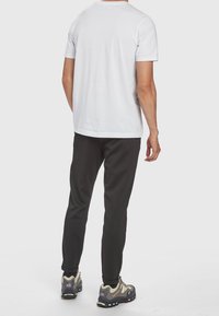 Man i vit kortärmad t-shirt, svarta byxor och grå vandringsskor, står vänd bort från kameran mot en ljus slät bakgrund.