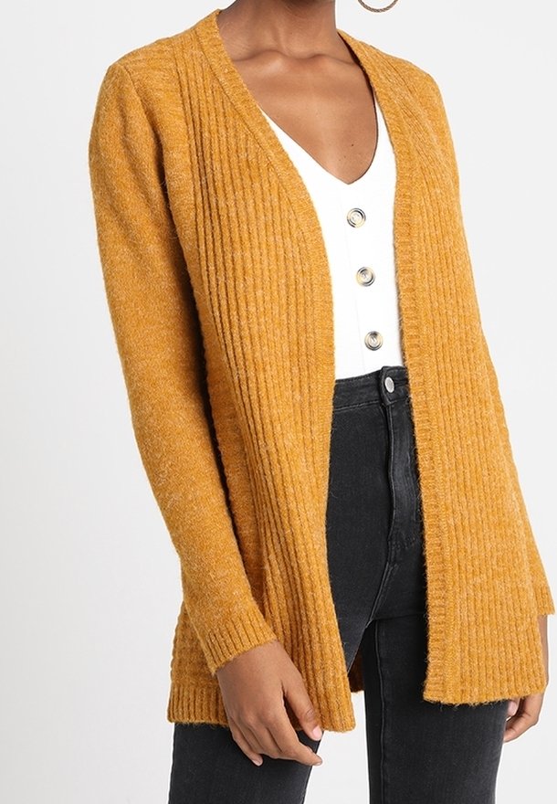 Femme portant un cardigan moutarde en maille côtelée, ouvert sur le devant, par-dessus un haut blanc boutonné et un jean taille haute noir, avec une boucle d'oreille visible.