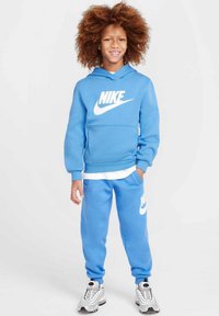 Sudadera azul de Nike con logo blanco y bolsillo tipo canguro, combinada con pantalones de chándal azules a juego que cuentan con un logo. Estilizada con zapatillas deportivas grises.