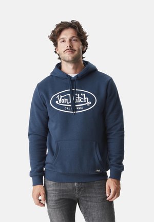 Homme aux cheveux ondulés portant un sweat à capuche Von Dutch bleu marine et un jean gris, debout devant un fond clair uni.
