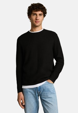Giovane uomo con capelli ricci che indossa un maglione nero a maglia sopra una camicia bianca, in piedi con una mano nella tasca dei jeans azzurri.