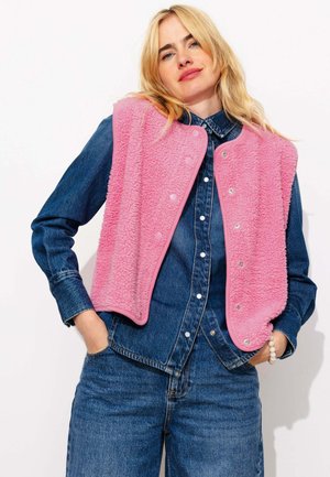Femme aux cheveux blonds portant un gilet rose duveteux par-dessus une veste en jean et un jean, posant les mains dans les poches devant un fond blanc.