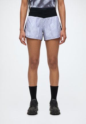 Un modèle se tient debout, vêtu d’un short de course clair à motifs avec une ceinture noire, de chaussettes noires et de chaussures de sport foncées.