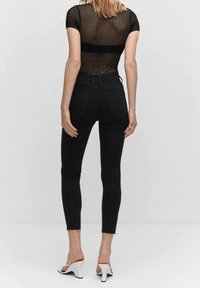 Jeans skinny negros de cintura alta con dobladillo deshilachado, combinados con una blusa de malla negra. Modelo usando sandalias de tacón blancas, de espaldas.