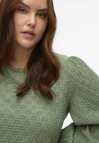 Blusa verde con texture e maniche a palloncino, caratterizzata da un motivo floreale e un collo rotondo, realizzata in un morbido tessuto intrecciato.