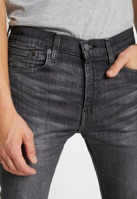 Se muestra la parte media de una persona que lleva unos jeans ajustados negros desgastados y una camiseta gris clara, con la mano derecha parcialmente en el bolsillo y la mano izquierda relajada a un lado.