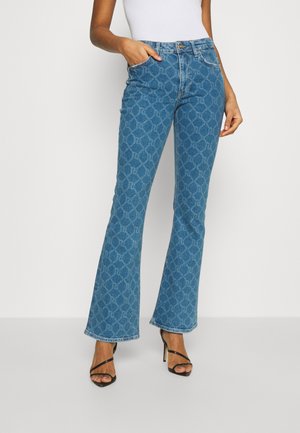 Žena v modrých džínech se vzorem a rozšířenými nohavicemi, bílém tílku bez rukávů a černých sandálech na vysokém podpatku, stojící s jednou rukou v kapse.