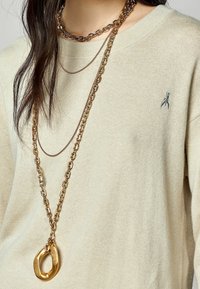 Collar de cadena dorada superpuesta sobre un suéter beige claro, con eslabones de diferentes tamaños y un prominente colgante ovalado.