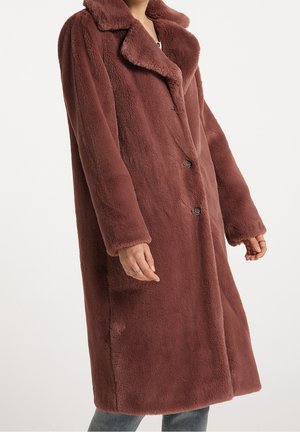 Femme portant un long manteau marron en fausse fourrure peluche boutonné sur un pantalon décontracté, debout devant un fond clair uni.