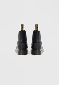 Fekete bőr bokacsizmák sima textúrával, masszív gumitalppal és kiemelkedő sárga húzófülekkel a hátsó részen. Kerek orrú kialakítás.