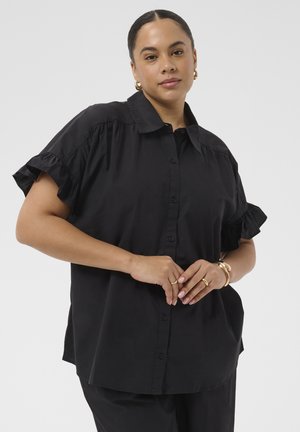 Femme aux cheveux tressés porte une chemise noire ample à boutons avec manches courtes à volants, ainsi que des boucles d’oreilles créoles dorées, des bagues et un bracelet.