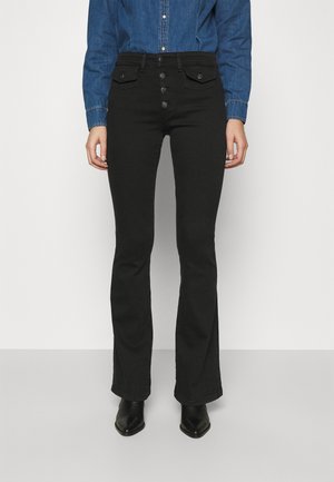 Personne portant un pantalon bootcut noir taille haute avec fermeture par boutons, des chaussures noires pointues et une chemise en denim bleu à manches longues, debout devant un fond uni.