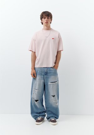 Fiatal férfi fehér falnak támaszkodik, halvány rózsaszín túlméretezett pólót, laza, szakadt kék farmert, lila sportcipőt és ezüst láncot visel.