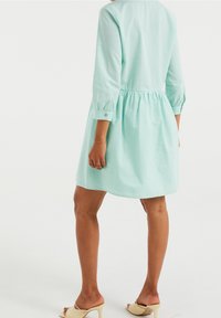 Mátová zelená šaty s třičtvrtinovými rukávy, nařaseným pasem a texturovanou tkaninou. Délka šatů sahá nad kolena. Spárováno se světle zbarvenými sandály.