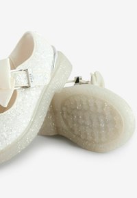 Scarpe bianche scintillanti con una lucente fibbia argentata, un grande accento a fiocco e una suola in gomma trasparente testurizzata con motivi esagonali.