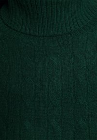 Pull vert foncé en maille torsadée avec un col montant côtelé, montrant des fibres de laine texturées et des motifs tressés verticaux.