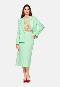 Mint green tailored blazer and matching pleated skirt, layered over a patterned top featuring floral designs. Red pointed-toe heels.