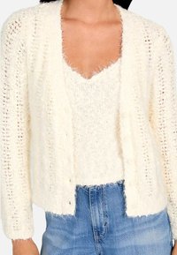 Cardigan alb pufos, cu deschidere frontală, mâneci lungi și un model texturat, purtat peste un pulover potrivit de același ton și blugi evazați.