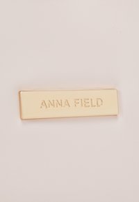 Etiqueta metálica rectangular dorada con el texto en relieve "ANNA FIELD", montada sobre una superficie lisa de color claro.