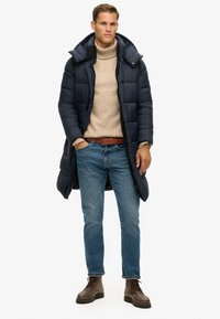 Marinen puffjakke med høj krave, beige strikket sweater, blå jeans, brun læderbælte og brune snøresko.