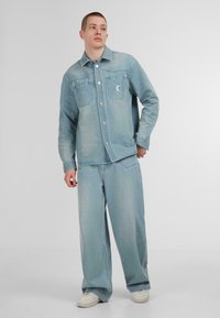 Lichtblauwe denim jas met twee borstzakken, knopen en een logo patch, gecombineerd met bijpassende wijde broek en witte sneakers.