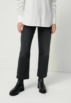 Personne portant un jean noir court, des bottes en cuir noires et une chemise blanche à manches longues, debout contre un mur uni.