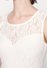 Femme portant une robe blanche sans manches en dentelle avec un motif floral et un panneau en dentelle transparente au niveau du décolleté.