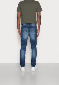 Jeans de mezclilla azul marino con corte slim, diseño de cinco bolsillos y una etiqueta de cuero marrón en la cintura trasera, combinados con una camiseta verde.