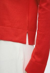 Haut rouge à manches longues avec fente latérale à l'ourlet porté sur un pantalon blanc, montrant la texture du tissu et les détails des coutures.