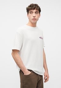 Νεαρός με σγουρά μαλλιά, φορώντας λευκό μπλουζάκι με μικρό λογότυπο στο στήθος και καφέ παντελόνι, στέκεται με το ένα χέρι στην τσέπη.