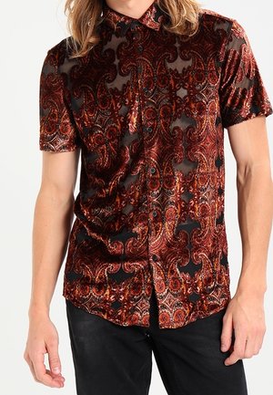 Koszula na guziki z krótkim rękawem w czerwono-czarno-pomarańczowy wzór paisley, noszona z czarnymi spodniami.