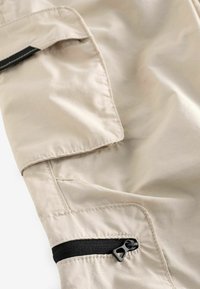 Pantalon cargo beige avec une texture lisse, poches latérales équipées de fermetures éclair noires et détails de couture, mettant l'accent sur la praticité et la fonctionnalité.