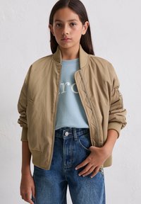 Beige Bomberjacke mit gerippten Bündchen und Kragen, kombiniert mit einem hellblauen Sweatshirt und hochtaillierten blauen Jeans.