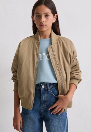 Beige bomberjack met geribde manchetten en kraag, gecombineerd met een lichtblauw sweatshirt en hoge blauwe spijkerbroek.