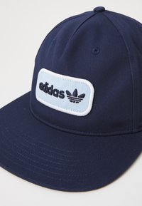 Námořnická modrá baseballová čepice s plochým kšiltem. Obsahuje světle modrou vyšívanou nášivku se „adidas“ a logem trefoil v černé barvě. Bavlněný materiál.