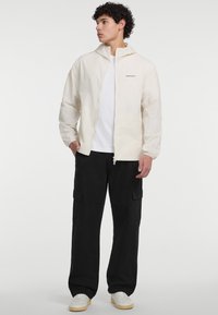 Jachetă albă ușoară cu glugă, închidere cu fermoar și un logo subtil pe piept, asociată cu pantaloni cargo negri și adidași albi.