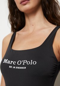Bañador negro con escote cuadrado, tirantes gruesos y texto blanco que dice "Marc O'Polo EST. EN ESTOCOLMO" en la parte frontal.