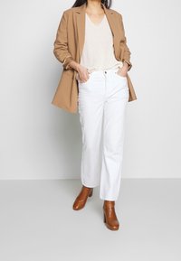 Brązowa marynarka, beżowy sweter z dekoltem w serek, białe proste spodnie oraz brązowe botki na obcasie. Czyste linie, neutralne kolory i teksturowany materiał.